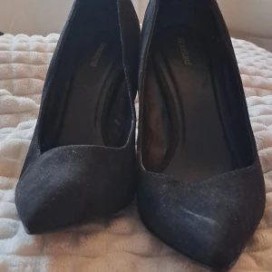 Svarta mocka pumps med snörning - Snygga svarta pumps från Graceland i mocka med spetsig tå och hög klack. Baksidan har cool snörning med guldfärgade detaljer som ger en unik touch. Perfekta för dig som vill sticka ut med stilrena och trendiga klackar.