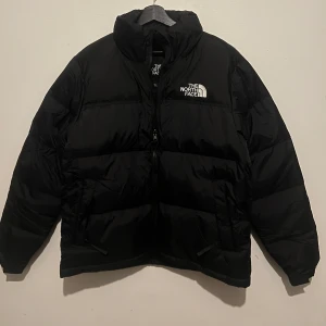 Svart pufferjacka från The North Face - Svart pufferjacka från The North Face med broderad logga på bröstet och ryggen. Jackan har hög krage, dragkedja framtill och två sidofickor. 700-fill märkning på ärmen. Perfekt för kalla dagar och har en clean, oversized vibe.