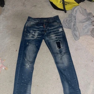Dsquared2 blåa jeans - Säljer ett par blå jeans från Dsquared2 med coola slitningar, hål och färgstänk. Jeansen har en klassisk femficksmodell, blå patch bak och Dsquared2-logga vid fickan. Materialet är bomull och polyester, passformen är rak och de har en modern streetkänsla.