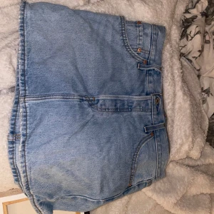 Levi's ljusblå jeanskjol - Säljer en ljusblå jeanskjol från Levi's med klassiska fem fickor, bälteshällor och knappstängning fram. Kjolen har rå kant nedtill och den ikoniska Levi's-lappen baktill. Perfekt för dig som gillar denim och vill ha en chill stil.