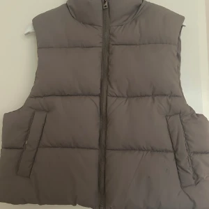 Beige/grå kort väst  - Fin vadderad väst med dragkedja framtill och två fickor på sidorna. Perfekt att bära över en hoodie eller tröja under kyligare dagar.