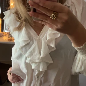 Vit blus med volanger - Säljer en vit blus med stora volanger längs v-ringningen. Blusen har knappar hela vägen och puffiga långärmar.