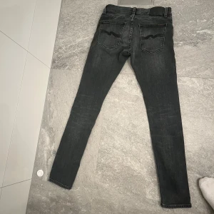 Svarta  jeans från Nudie - Säljer ett par svarta skinny jeans från Nudie med klassiska femficksdetaljer och Nudie-loggan på bakfickorna. Jeansen har smal passform och är tillverkade i stretchig denim för extra komfort. Perfekta för dig som gillar en  look.
