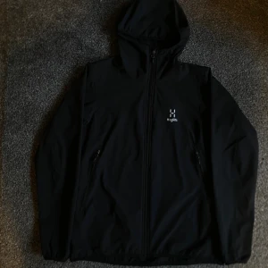 Svart vindjacka från Haglöfs - Svart vindjacka från Haglöfs med huva och hel dragkedja framtill. Jackan har två sidofickor med dragkedja och en stilren logga på bröstet. Perfekt för blåsiga dagar och har en clean, sportig look som funkar till streetwear.