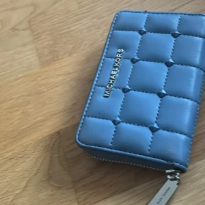 Blå quiltad plånbok Michael Kors - Snygg blå plånbok från Michael Kors i quiltad design med silvrig dragkedja och logga. Plånboken har flera fack för kort och sedlar samt en praktisk ficka på baksidan. Tillverkad i skinn med stilren look.