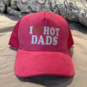 Rosa keps med texten I ❤️ HOT DADS - Säljer en rosa truckerkeps med vit texten 'I ❤️ HOT DADS' framtill. Kepsen har mesh på sidorna och bak, samt justerbar snapback för perfekt passform. Perfekt statement-piece för dig som vill sticka ut.