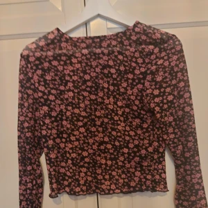 Svart meshblus med rosa blommor - ⚠️ Skriv till mig innan köp!⚠️Säljer en svart meshblus med lång ärm och rosa blommigt mönster. Toppen är lätt transparent och har en croppad passform med vågig kant nedtill. Perfekt för dig som gillar att sticka ut med coola prints och mjuka material.