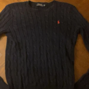 Mörkblå kabelstickad tröja Polo Ralph Lauren - Kabelstickad mörkblå tröja från Polo Ralph Lauren med klassisk röd broderad logga på bröstet. Tröjan har rund halsringning och långa ärmar, perfekt för lager-på-lager under höst och vinter. Helt ny aldrig använd. 