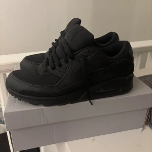 Nike Air Max 90 helsvarta sneakers - Nike Air Max 90 sneakers i helsvart färg med klassisk siluett och synlig Air-enhet i sulan. Skorna har snörning, rund tå och platt sula. Ovandelen är en mix av skinn och syntetmaterial med meshpaneler för extra komfort. Perfekt för dig som gillar stilrena och sportiga sneakers. Orginal lådan är borttappad! Kommer istället i en air force 1 låda!