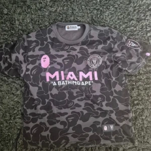 Svart camo fotbollströja BAPE x Inter Miami - Unik fotbollströja från BAPE x Inter Miami i svart och grått camouflagemönster med rosa tryck. Kortärmad modell med rund hals, tillverkad i mjukt bomullsmaterial. Framsidan har MIAMI-logga och BAPE-ikon, baksidan har stort rosa 93-tryck och BAPE-text.