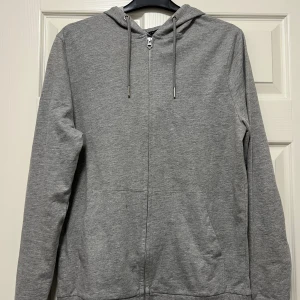 Grå hoodie från ASOS DESIGN - En stilren grå hoodie från ASOS DESIGN med dragkedja framtill, huva med snörning och två fickor. Tillverkad i mjuk bomullsblandning för extra komfort. Perfekt för chill dagar eller när du vill ha en enkel och clean look. Liten fläck finns, men syns knappt. Se sista bild. Sparsamt använd, så fint skick!