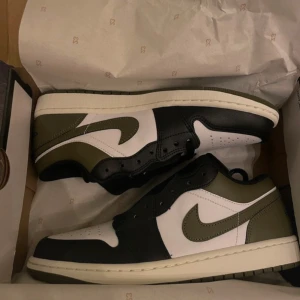 Nike Air Jordan 1 Low Olive/Black - Nike Air Jordan 1 Low i olivgrön, svart och vit färg. Snygg design med klassisk Jordan-logga på hälen och ikonisk Swoosh på sidan. Låg modell med snörning och platt sula. Materialet är skinn och syntet, perfekt för dig som gillar streetwear och sneakers med attityd.