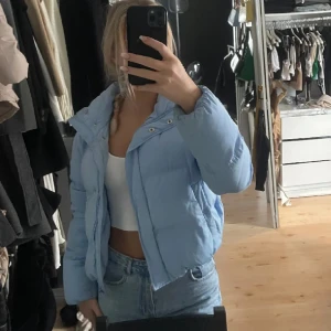 Ljusblå pufferjacka med huva - Finns lite sminknärken vid kragen. Säljer en ljusblå pufferjacka med huva och snygga detaljer som tryckknappar framtill. Jackan är croppad och har en loose passform, perfekt för att ge en chill vibe till din outfit. Materialet är mjukt och syntetiskt, vilket gör den både lätt och varm. 