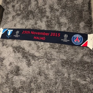 Champions League halsduk Malmö PSG - Unik halsduk från matchen Malmö FF vs Paris Saint-Germain den 25 november 2015. Ena sidan är marinblå med röda och vita detaljer, den andra sidan är röd med svarta tryck. Båda sidor har klubbmärken och Champions League-logga, samt vita fransar i ändarna. Perfekt för fotbollsfans! Använd en gång! Limited edition