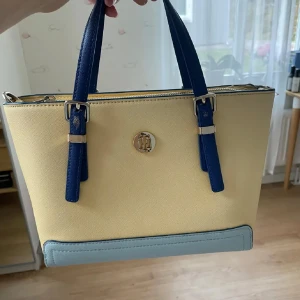 Handväska från Tommy Hilfiger i pastell - Snygg handväska från Tommy Hilfiger i ljusgul och ljusblå färg med mörkblå handtag. Väskan har ett stilrent TH-emblem i guld på framsidan och dragkedja upptill. Materialet är syntet med slät finish och detaljer i metall. Perfekt storlek för vardagsgrejer.