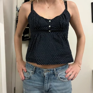 Mörkblå prickig linnetopp med knyt - Säljer en mörkblå linnetopp med vita prickar och smala axelband. Toppen har tre dekorativa knappar framtill, lätt rynkade detaljer vid bysten och ett sött knyt i ryggen. Materialet känns som bomull och toppen är croppad med en loose passform. Märket är Brandy Melville och storlek S.