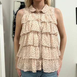 Prickig volangblus i ljusrosa - Säljer en söt blus i ljusrosa med svarta prickar och flera lager volanger framtill. Blusen är ärmlös, har en rund krage och knäppning hela vägen fram. Materialet är lätt och luftigt, perfekt för varmare dagar. Superfin till jeans eller kjol! Märket är Lindex och storlek S (170).