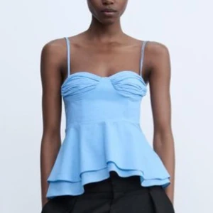 Ljusblå volang topp med smala axelband - Supersöt ljusblå topp från Zara med smala axelband och volang nedtill. Kuporna har snygga rynkdetaljer och baksidan har knytband för extra passform. Perfekt för dig som gillar en feminin och trendig look.
