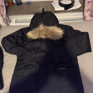 Svart parkas från Canada Goose - Säljer en svart parkas från Canada Goose med huva och snygg pälsdetalj. Jackan har två stora fickor framtill och klassisk logga på ärmen. Perfekt för kalla dagar med sin varma och rejäla design. Stängs med dragkedja och har en loose passform.