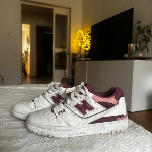 New Balance 550 sneakers vinröd/rosa - Säljer ett par New Balance 550 sneakers i vitt med vinröda och rosa detaljer. Skorna har snörning, perforerade paneler och platt sula. Ikonisk N-logga på sidan och bekväm mesh vid kragen. Perfekt för dig som gillar retrostil och streetwear.