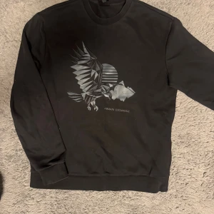 Svart sweatshirt Armani Exchange S - Svart sweatshirt från Armani Exchange i storlek S med ett stilrent geometriskt örnmotiv i silver på bröstet. Tröjan har rund hals, långa ärmar och ribbade muddar. Perfekt för dig som gillar grafiska prints och vill ha en cool streetstyle-look.