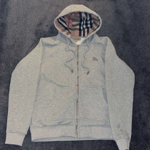 Grå hoodie från Burberry med dragkedja - Grå hoodie från Burberry med klassiskt rutigt foder i huvan och insidan. Tröjan har snörning vid huvan, broderad Burberry-logga på bröstet och praktiska fickor framtill. Materialet är mjuk bomull och polyester, perfekt för chill dagar. Pris kan diskuteras!!!