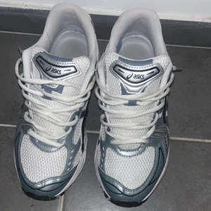 Vita och grå Asics sneakers - Säljer ett par fräscha Asics sneakers i vitt och grått med klassisk mesh och syntetpaneler. Skorna har snörning, rund tå och en chunky sula med GEL-dämpning för extra komfort. Perfekta för dig som gillar sportig och trendig stil.