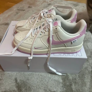 Nike Air Force 1 Low vit/rosa - Nike Air Force 1 Low sneakers i vit och ljusrosa med klassisk snörning och rund tå. Ovandel i skinn med rosa swoosh och detaljer, samt rosa yttersula. Mjuk mesh på insidan för extra komfort. Perfekt för dig som gillar clean och färgglad stil.