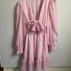 Rosa volangklänning med knyt - Supersöt ljusrosa kortklänning med volanger, långärmade puffärmar och smock i midjan. Djup v-ringning med knyt fram och volangdetaljer vid ärmslut och kjol. Tunt, flowigt material som ger en romantisk vibe.