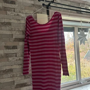 Randig röd och rosa långärmad klänning - Säljer en cool långärmad klänning med breda horisontella ränder i rött och rosa. Klänningen har en rundad, djup urringning både fram och bak och är i en mjuk, tunn bomullskvalitet. Passformen är normal och längden går till strax ovanför knäna. Perfekt för dig som gillar färg och mönster!