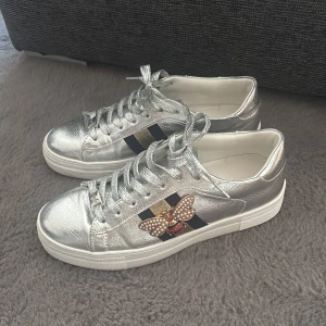 Silvriga sneakers med pärlbi - Snygga sneakers i metallic-silver med svarta och guldiga ränder på sidorna samt en cool detalj i form av ett pärlbi. Perfekt för dig som vill sticka ut! Storlek 37, mycket bra skick, endast använda ett fåtal gånger. Kan både skickas och hämtas upp!