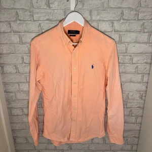 Ralph Lauren skjorta - Persikofärgad skjorta från Polo Ralph Lauren i slim fit med klassisk button down-krage och blå broderad logga på bröstet. Skjortan är tillverkad i 100% bomull och har långa ärmar samt knäppning framtill. Hör av dig vid intresse 🙌