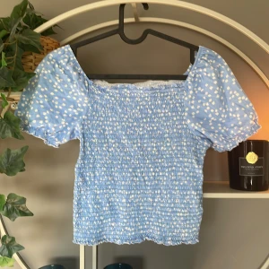 Blommig blå smockad topp KappAhl - Supersöt ljusblå topp från KappAhl med vita småblommor. Toppen har smockad kropp, volangkant upptill och nertill samt puffiga korta ärmar. Perfekt för en somrig look med romantiska vibes. Korsettliknande med puffiga armar gör det riktigt snyggt. Säljer för att den tyvärr är för liten 