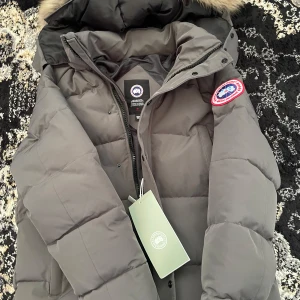  Canada Goose  - Säljer en grå dunjacka från Canada Goose med klassisk patch på ärmen och pälskantad huva. Jackan är quiltad, har dragkedja och tryckknappar framtill. Perfekt för kalla vinterdagar och har en clean, stilren look.