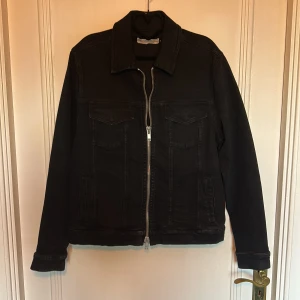 Svart jeansjacka med dragkedja - Säljer en svart jeansjacka från The Blue Uniform med klassisk krage och silverfärgad dragkedja framtill. Jackan har två bröstfickor med lock och knappar samt raka ärmar. Perfekt för dig som gillar en stilren och enkel look.