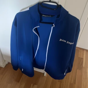 Blå Palm Angels ziptröja med vita ränder - Säljer en blå ziptröja från Palm Angels med vita ränder längs ärmarna och vit dragkedja. Tröjan har logga på bröstet och är tillverkad i polyester. Perfekt för dig som gillar streetwear och sportig stil.