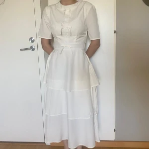 Vit långklänning med knyt och volanger - Superfin vit långklänning med flera lager volanger i kjolen och tre söta knyt framtill. Klänningen har halvkort ärm och rund halsringning. Materialet känns lätt och flowy, perfekt för varma dagar. Dragkedja i ryggen för enkel påklädning.