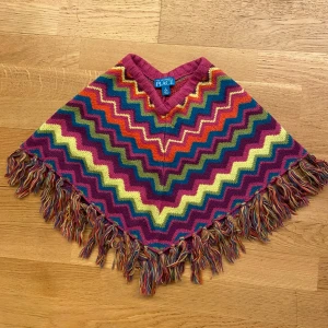 Färgglad zigzag poncho - Poncho i regnbågens färger från The Children's Place med coolt zigzag-mönster och fransar längs kanten. Stickad i 100% akryl och har en skön, loose passform. Perfekt för att liva upp höst- och vinterlooken!