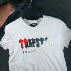 Vit Trapstar t-shirt med broderad logga - Vit t-shirt från Trapstar i bomull med broderad logga i rött, svart, blått och vitt framtill. Klassisk passform med rund hals och korta ärmar. Snygg streetwear-vibe och tydlig branding framtill.
