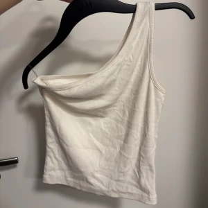 Vit ribbad one shoulder topp - Säljer en vit ribbad topp med one shoulder-design. Toppen är ärmlös och har en tight passform som framhäver axlarna. Perfekt för dig som gillar minimalistisk och trendig stil.