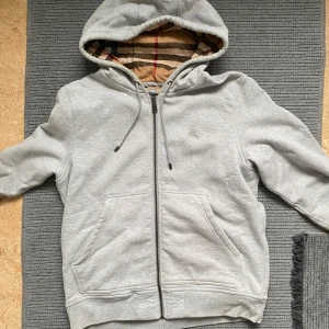 Burberry tröja med dragkedja - Snygg grå hoodie från Burberry med klassiskt rutigt foder i huvan. Tröjan har dragkedja, snörning i huvan och två stora fickor framtill.  Den är helt ny utan etikett, inga skador!