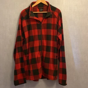 Röd och svart fleecejacka från Everest - Rutig fleecejacka från Everest i rött och svart. Jackan har hel dragkedja framtill, två fickor med dragkedja och hög krage. Perfekt för kyliga dagar och enkel att slänga på sig över en t-shirt. Mjuk och skön känsla.