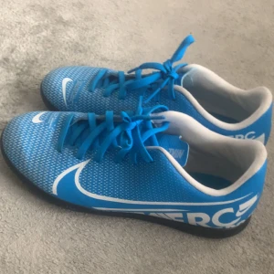 Blå Nike Mercurial fotbollsskor - Säljer ett par blå Nike Mercurial fotbollsskor med vit logga och snörning. Skorna har en platt sula med grepp, perfekt för inomhusspel. Ovandelen är i syntetmaterial och har ett modernt, sportigt mönster. Snygga och lätta skor för dig som älskar fotboll.