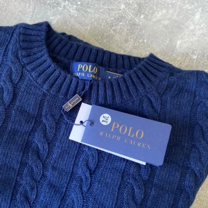 Marinblå kabelstickad tröja Polo Ralph Lauren - Kabelstickad marinblå tröja från Polo Ralph Lauren med rund halsringning och klassisk röd logga broderad på bröstet. Tröjan är stickad i mjuk bomull och har ribbade muddar vid ärmslut och nederkant. Perfekt för en stilren och tidlös look.