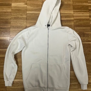 Vit hoodie med dragkedja - Snygg vit hoodie med hel dragkedja och huva. Tröjan har två stora fickor framtill och ribbade muddar vid ärmslut och nederkant. Perfekt för lager-på-lager och chill dagar. Enkel och stilren design som passar till det mesta.
