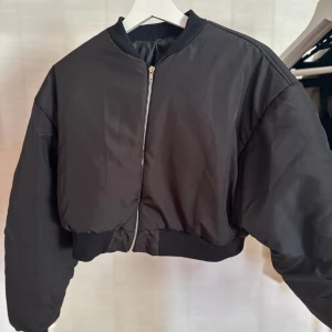 Svart croppad bomberjacka - Säljer en svart croppad bomberjacka med dragkedja framtill och ribbade muddar vid krage och midja. Jackan har en loose passform och är perfekt för dig som vill ha en stilren och trendig look. Materialet är syntet och jackan har långa ärmar.