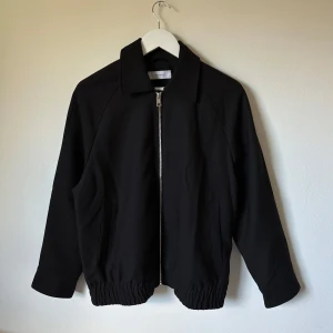 Svart skjortjacka från Reserved - Svart skjortjacka från Reserved med klassisk krage, dragkedja framtill och elastisk midja. Jackan är tillverkad i en mix av polyester, viskos och elastan, vilket ger en mjuk och följsam känsla. Perfekt för dig som gillar stilren och enkel design. 3Q19