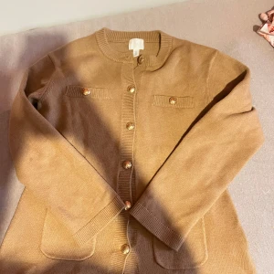 Beige kofta med guldknappar - Snygg beige kofta från H&M med guldiga knappar framtill och dekorativa fickor på bröstet. Långärmad och ribbade muddar vid ärmslut och nederkant. Perfekt för lager-på-lager och enkel att styla till olika outfits.