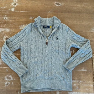 Grå kabelstickad tröja Polo Ralph Lauren - Kabelstickad grå tröja från Polo Ralph Lauren med halv dragkedja och hög krage. Klassisk broderad logga på bröstet. Tröjan är ribbad vid ärmslut och nederkant och har långärmad passform. Perfekt för lager-på-lager under kyliga dagar. Är endast använd ett visst antal gånger pga storlek. Priset är ej hugget i sten.