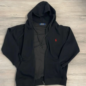 Svart hoodie från Polo Ralph Lauren M (ops passar endast S inte för M) - Svart Polo Ralph Lauren hoodie med röd broderad logga på bröstet. Tröjan har huva med snörning, dragkedja framtill och två fickor. Perfekt för en chill och stilren look, dock passar den mer som S än M, därför rekommenderar jag att du inte köper varan om du har M i storlek. Samt har varan aldrig används och därmed i perfekt skick.